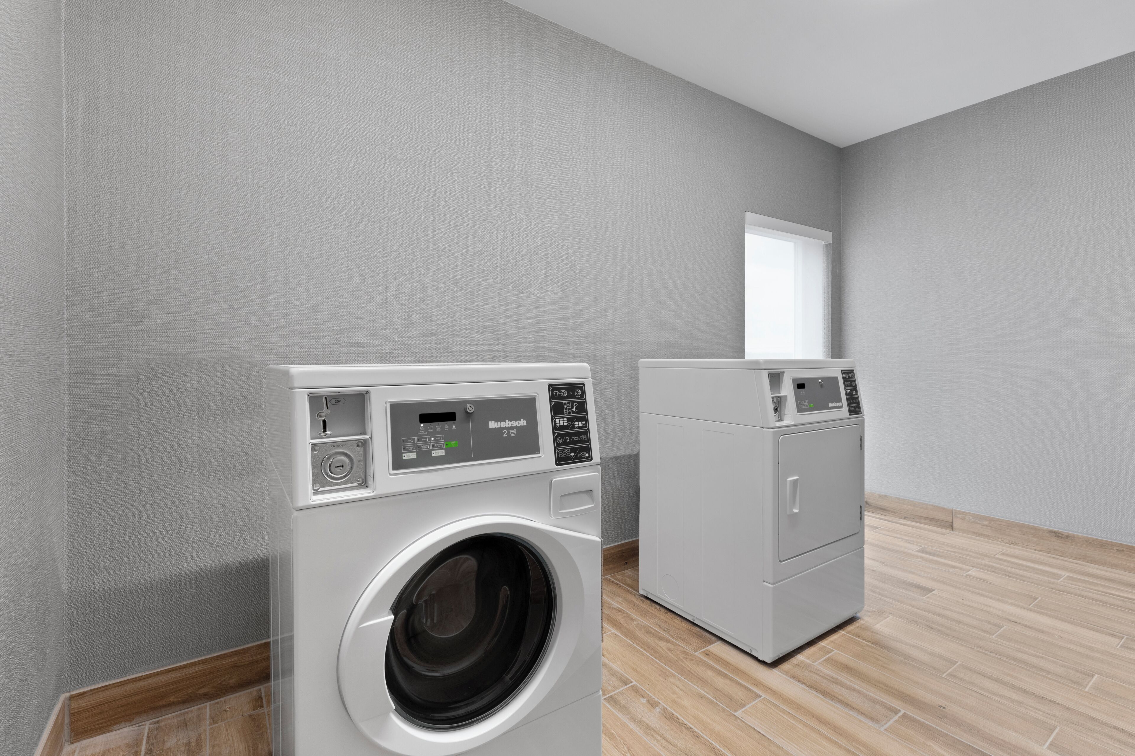A laundry room with two front-loading washing machines and a dryer, a window, and a wooden floor.