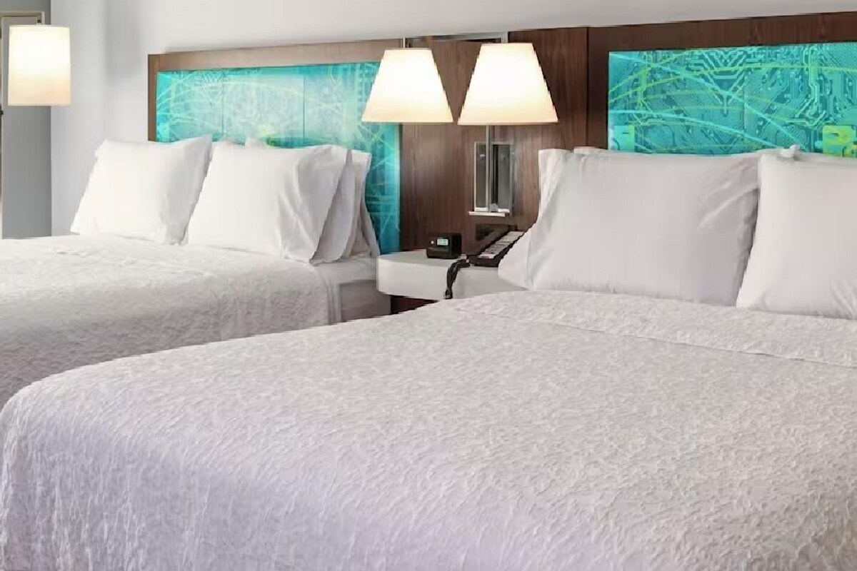 Habitación de hotel con dos camas, una mesita de noche, una lámpara y un panel con diseño de circuito electrónico.