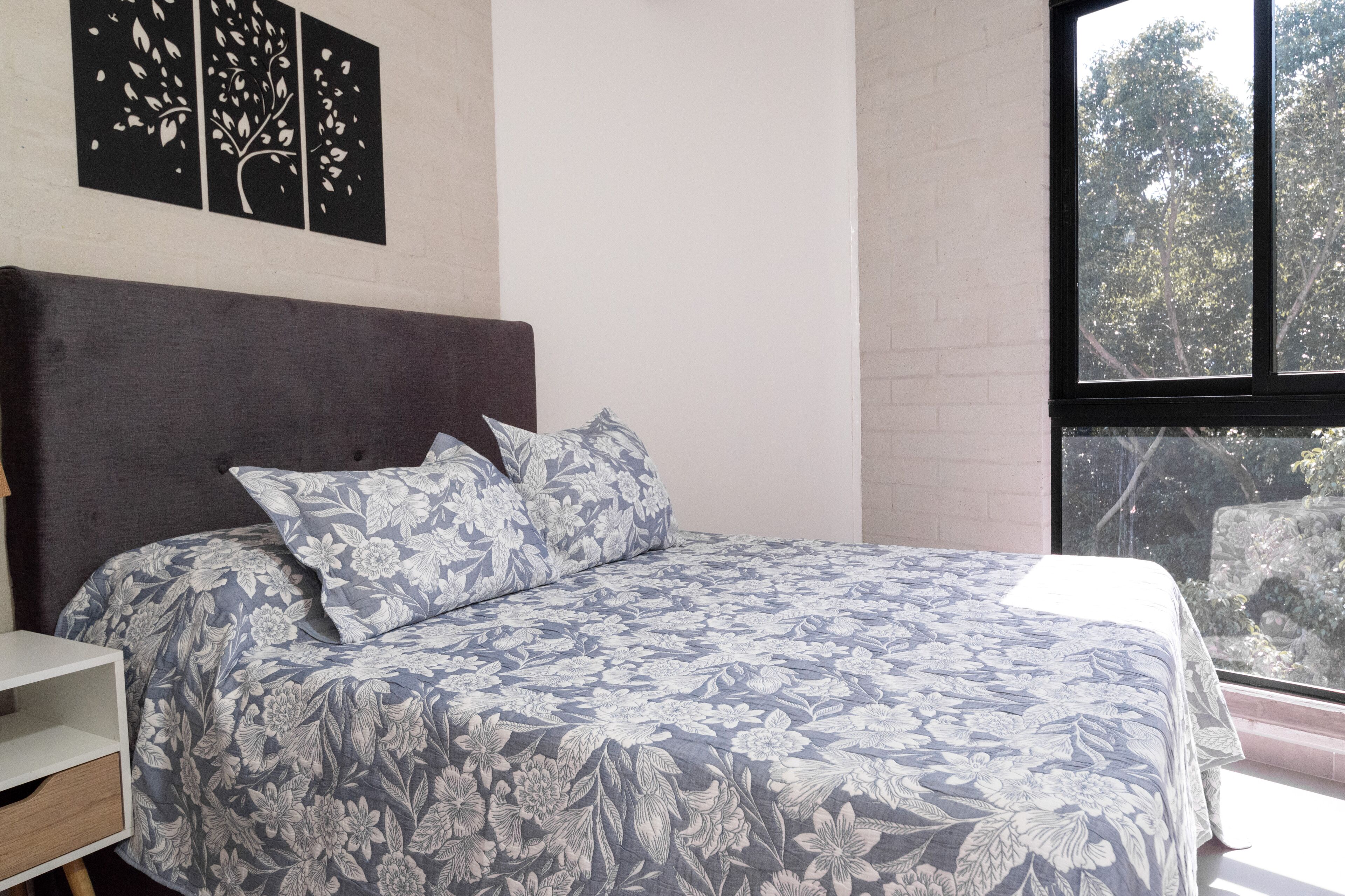 A bedroom with a bed, a nightstand, a window with a view of trees, and a wall with a tree decal.