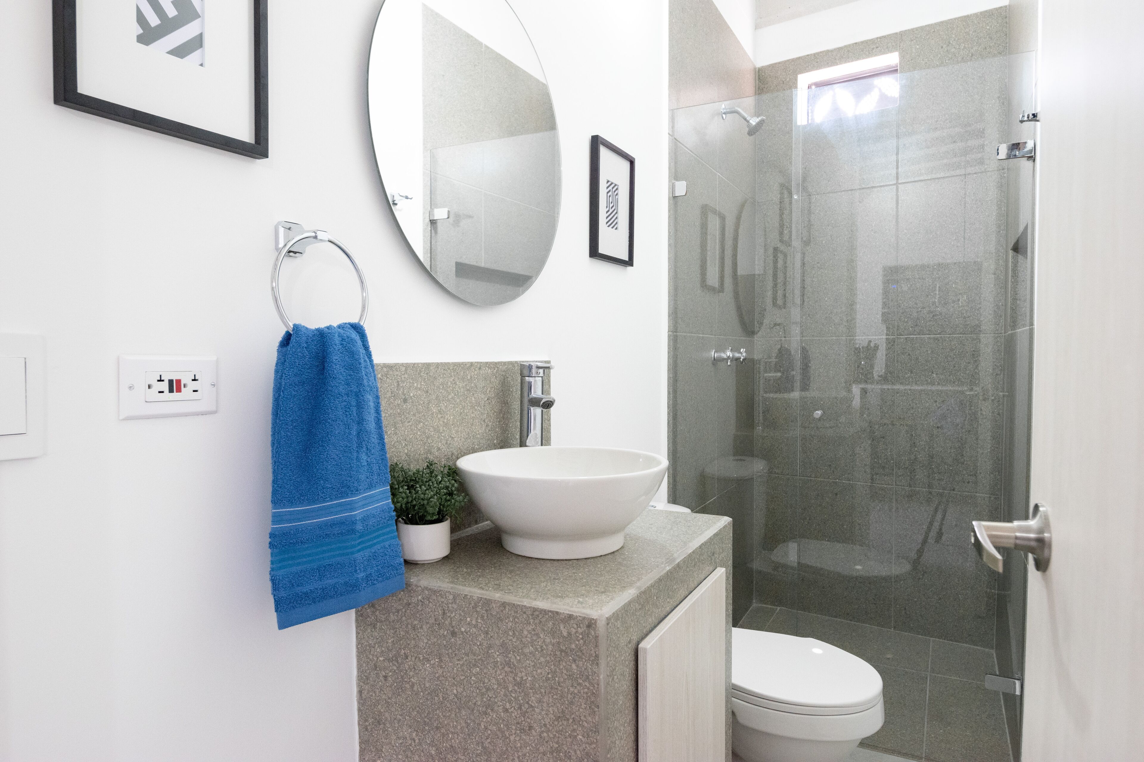 Un baño con un lavabo blanco, un espejo redondo, una toalla azul, una pequeña planta en maceta y una zona de ducha con paredes de vidrio.