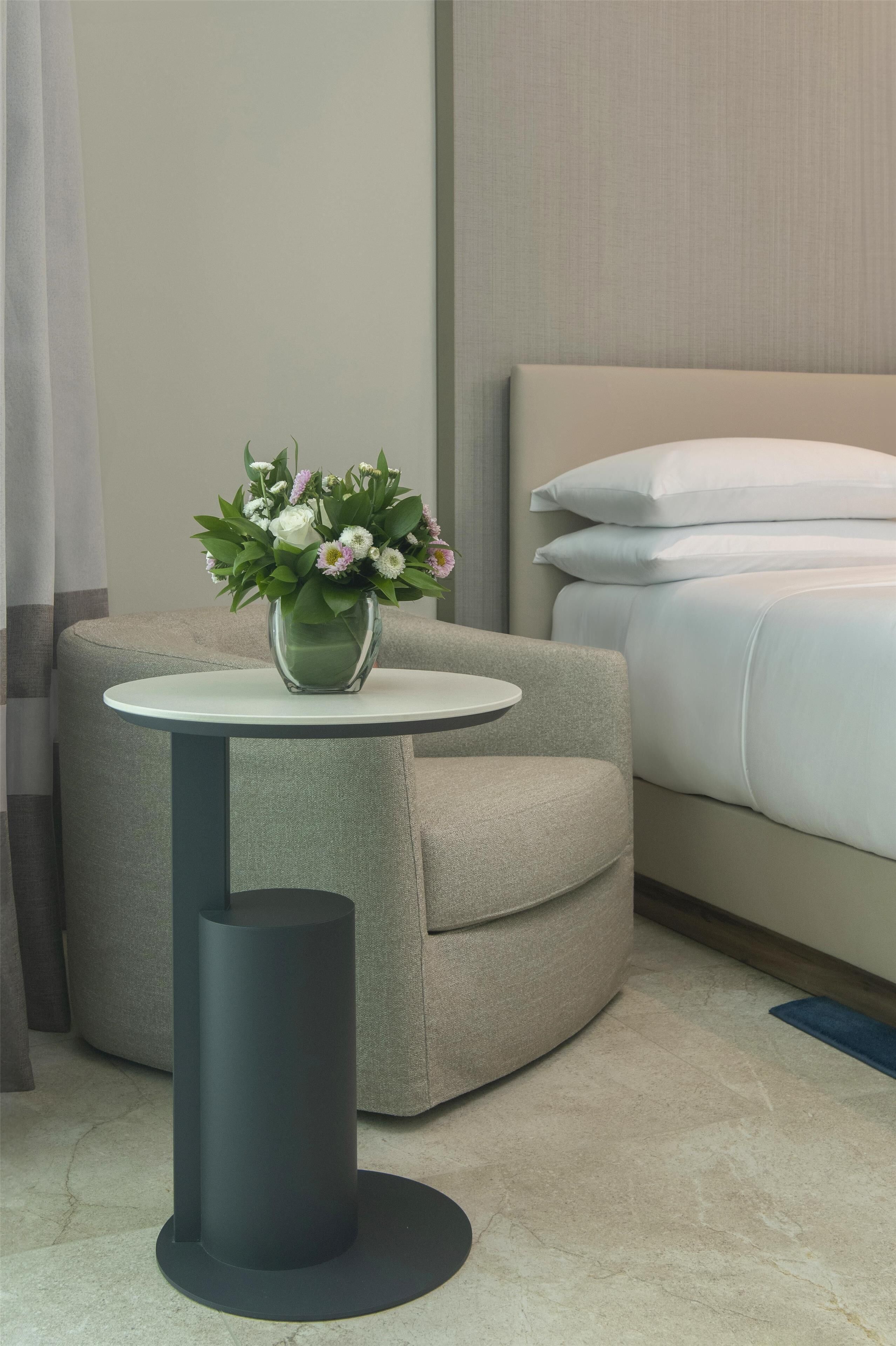 A modern hotel room with a grey sofa, a small round table with a vase of flowers, and a bed with white bedding.