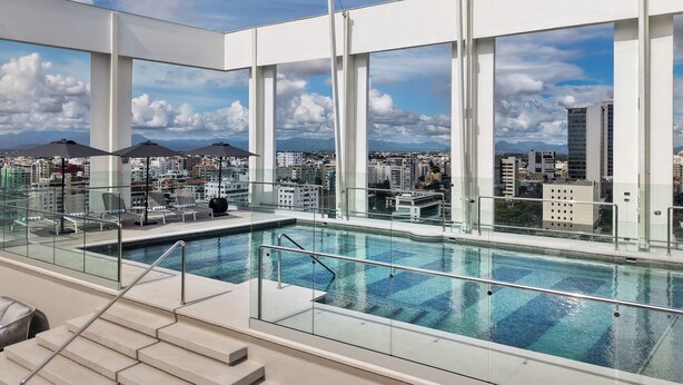 A rooftop pool with glass railings, a view of the cityscape, and a clear sky.