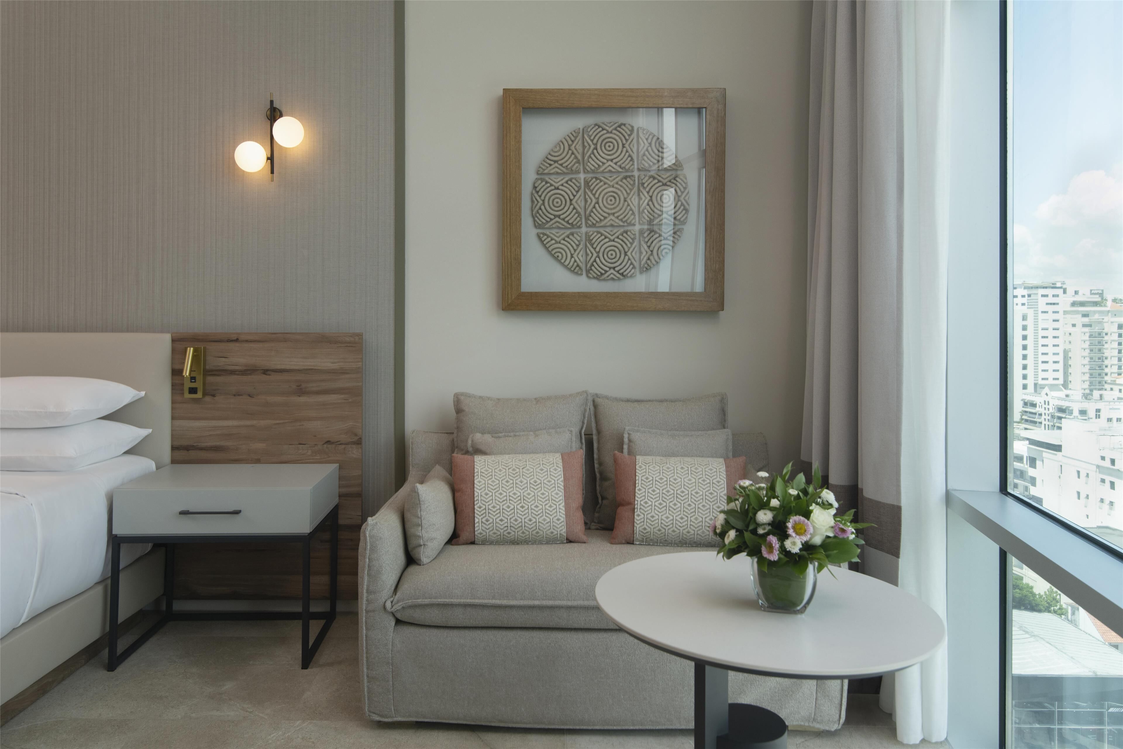 Una habitación de hotel moderna con una cama, un sofá, una mesita y un amplio ventanal con vistas a la ciudad.