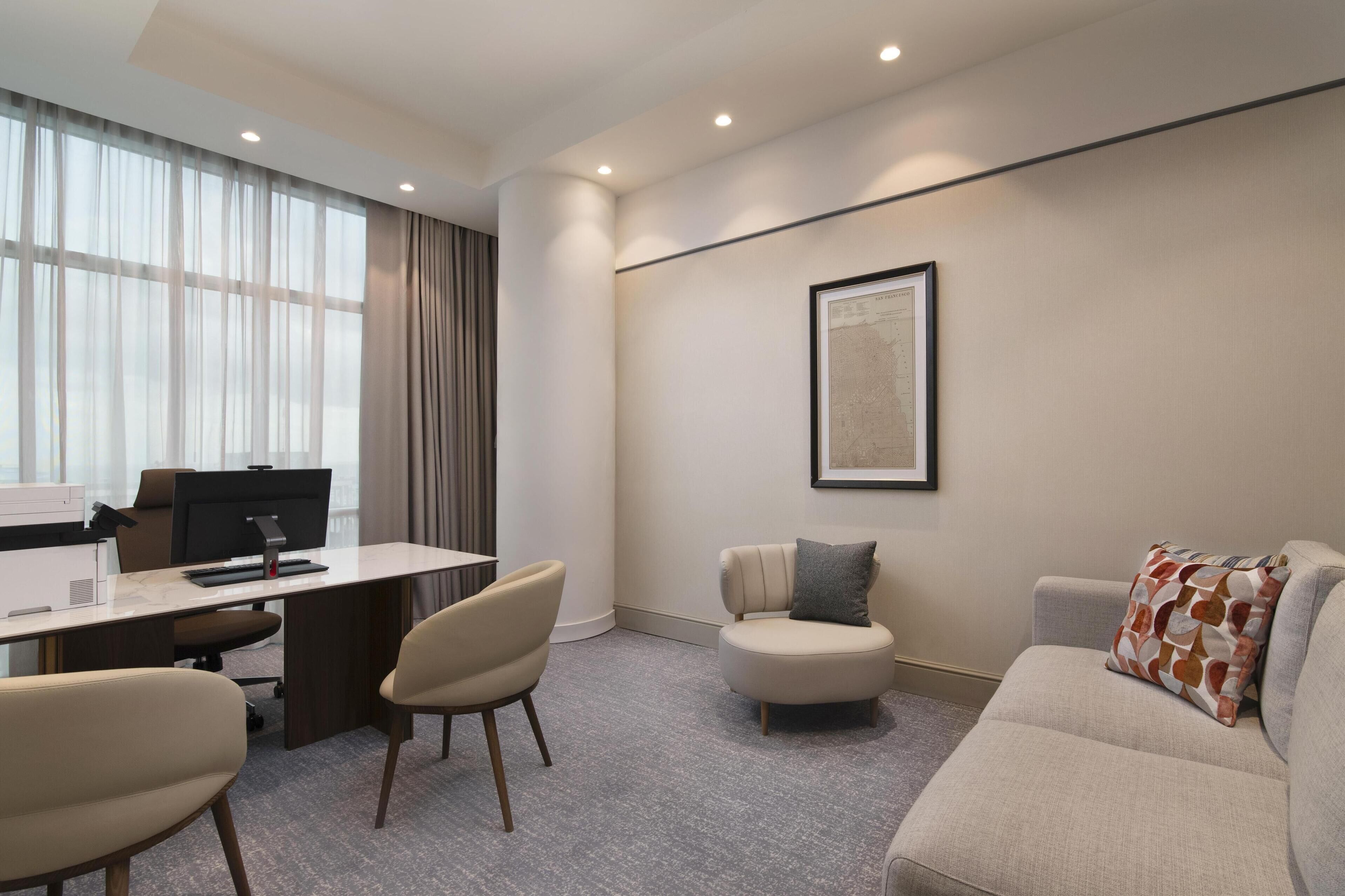 Een moderne hotelkamer met een bureau, twee stoelen, een bank en een groot raam met gordijnen.