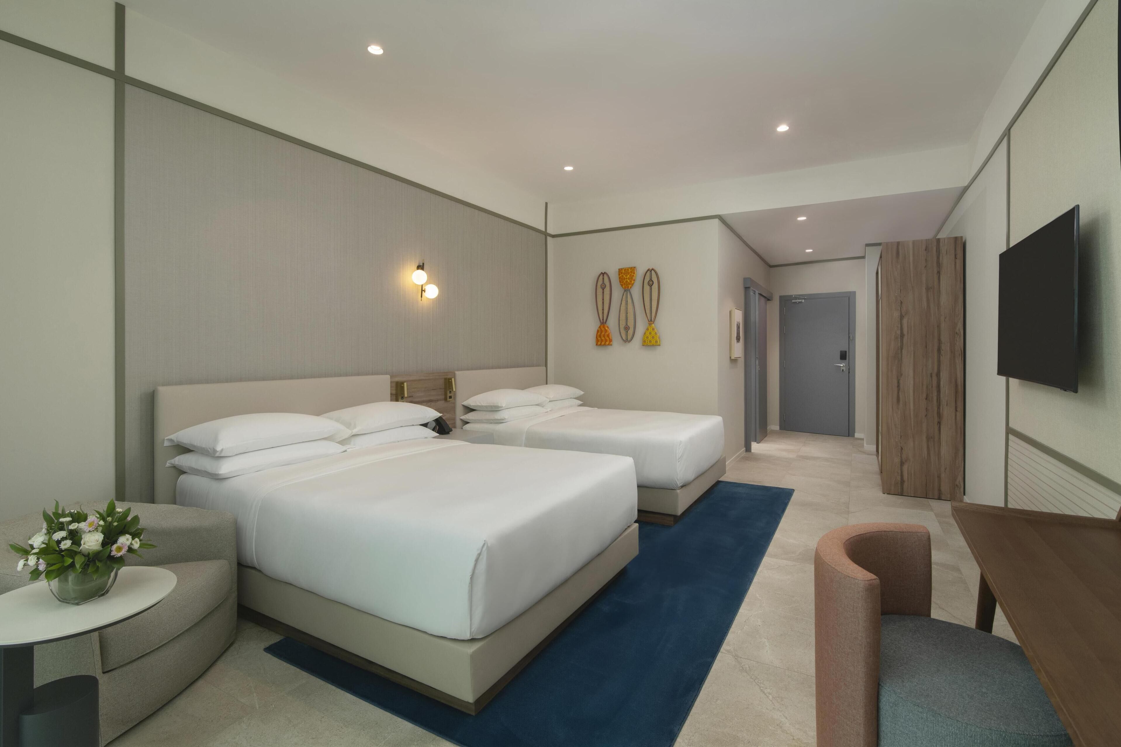 A modern hotel room with two beds, a flat-screen TV, a small round table with a vase of flowers, and a wooden desk.