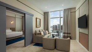 A modern hotel room with a bed, sofa, and armchair, a TV, and a large window with a city view.