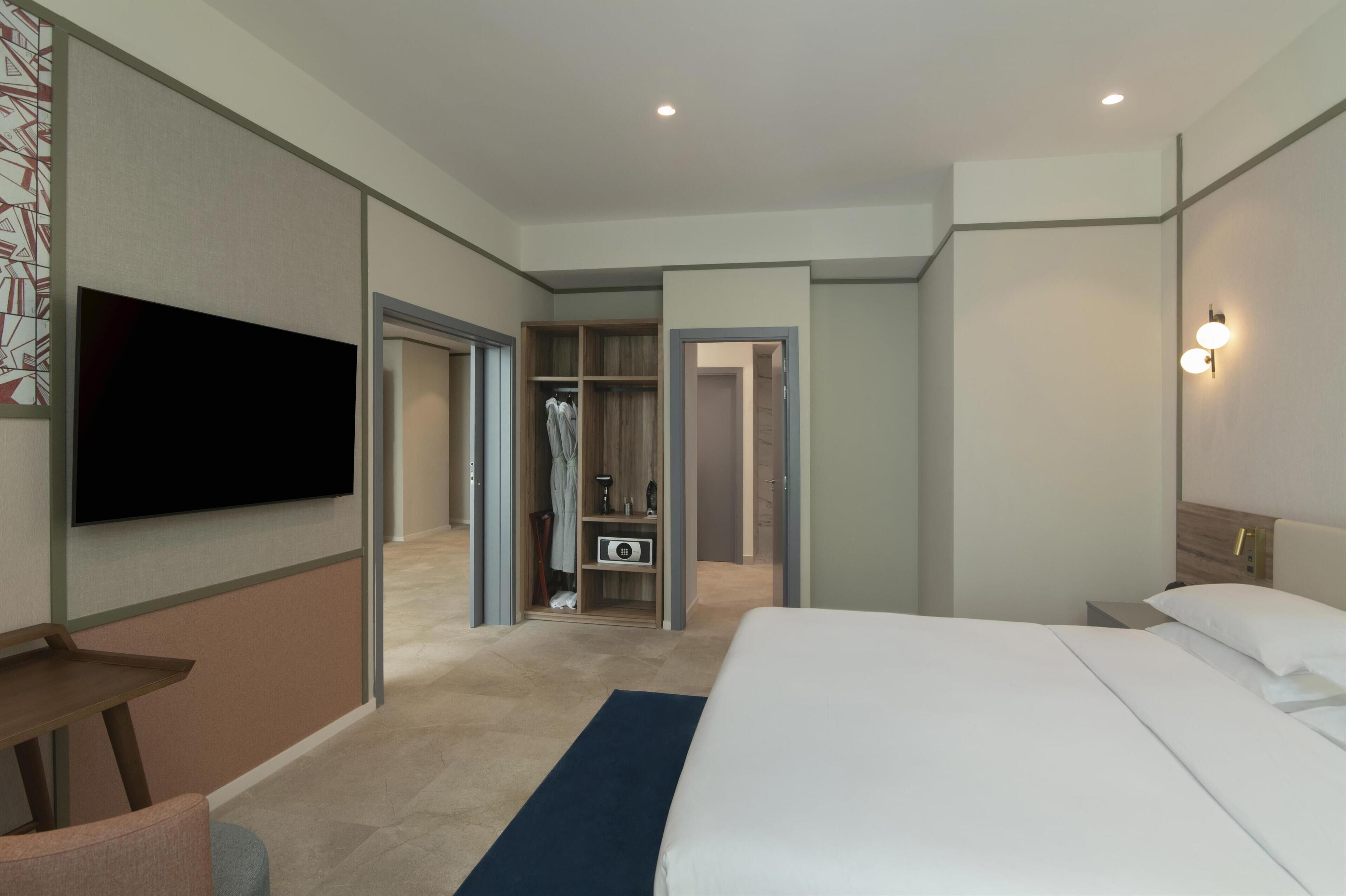 Una habitación de hotel moderna con una cama grande, un televisor de pantalla plana en la pared, un vestidor amplio y una mesita de noche.