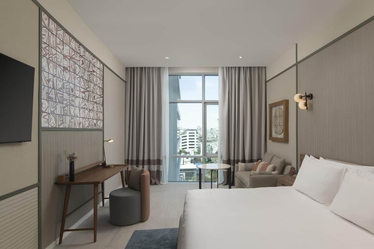 A modern hotel room with a large bed, a desk, a chair, and a small table. There is a large window with curtains, a wall-mounted TV, and a framed picture.