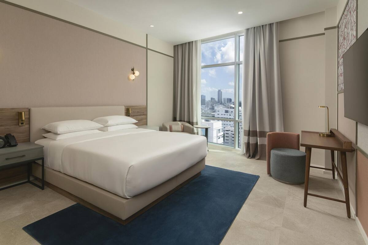 A modern hotel room with a large bed, a desk, a chair, a small round ottoman, and a TV.