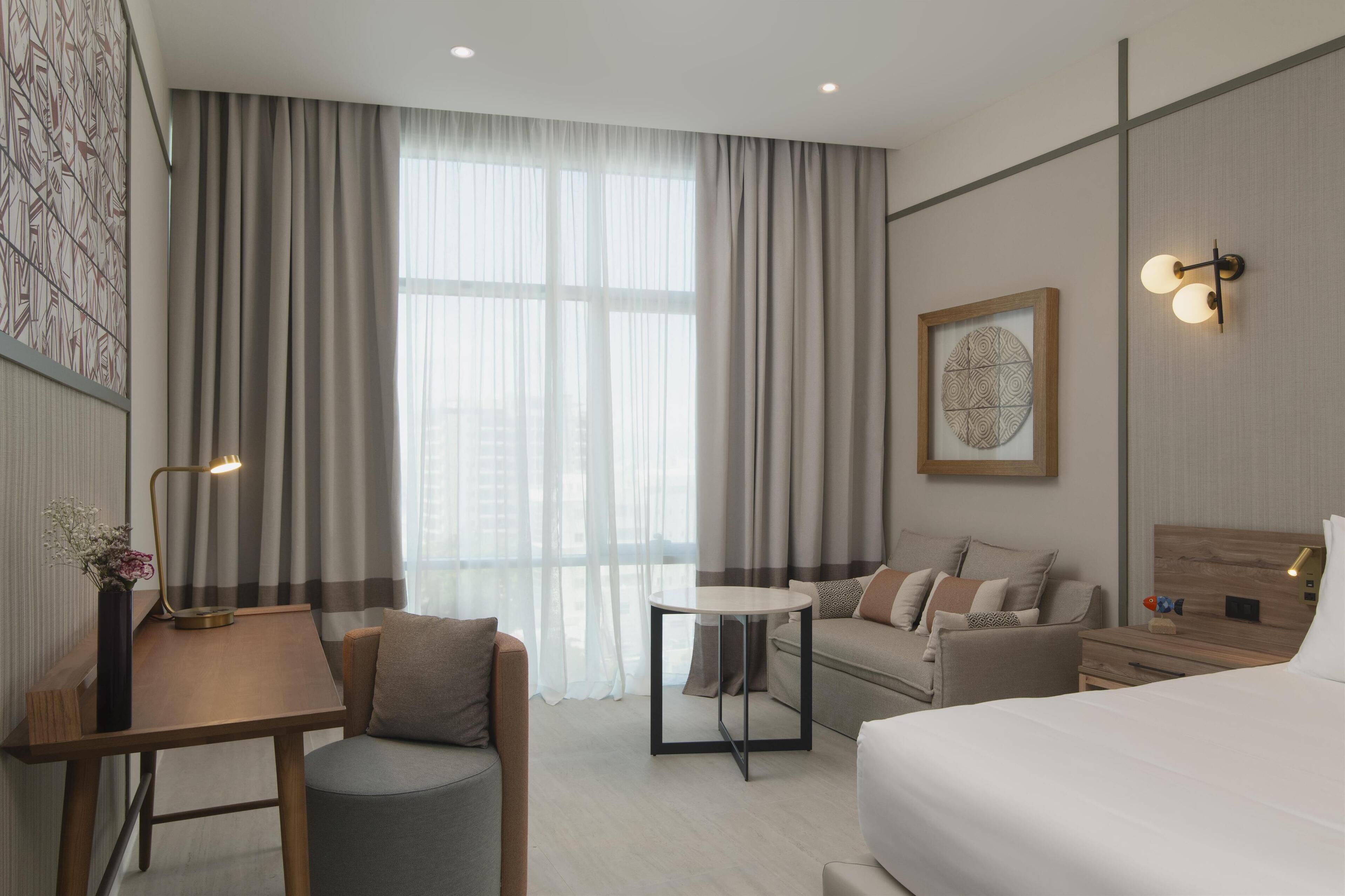Ett modernt hotellrum med en säng, en soffa, ett litet bord och ett skrivbord. Det finns ett stort fönster med gardiner, en vägglampa och en inramad tavla.