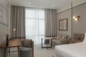 Una habitación de hotel moderna con una cama, un sofá, una mesita y un escritorio. Cuenta con un amplio ventanal con cortinas, una lámpara de pared y un cuadro enmarcado.