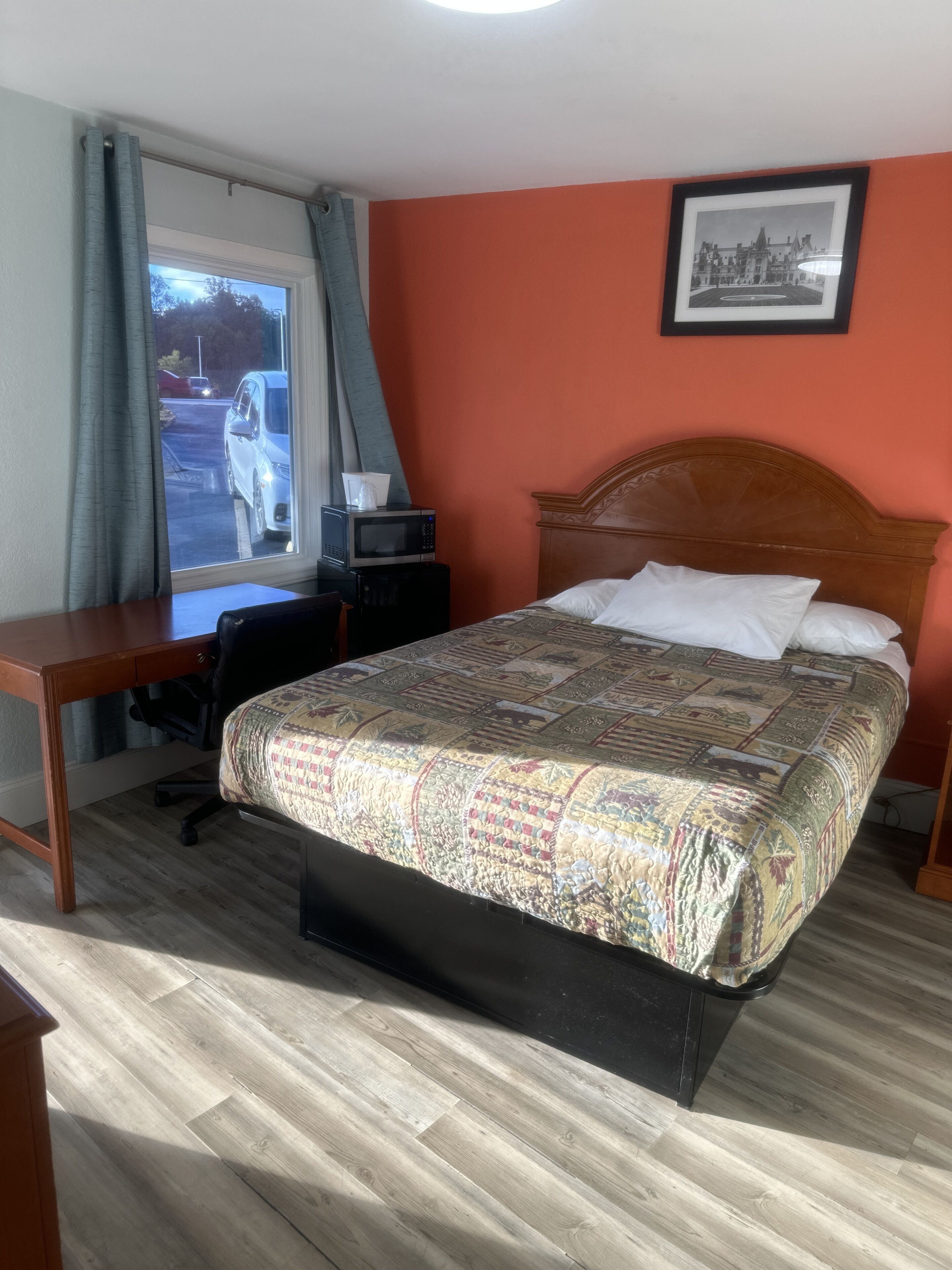 Quarto de hotel com cama, escrivaninha, cadeira, janela com cortinas e um quadro na parede.