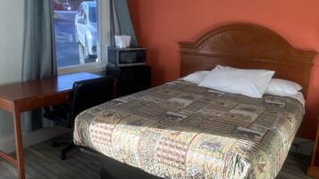 Quarto de hotel com cama, escrivaninha, cadeira, janela com cortinas e um quadro na parede.