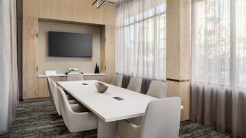 A conference room with a long table, six chairs, a flat-screen TV mounted on the wall, and large windows with sheer curtains.