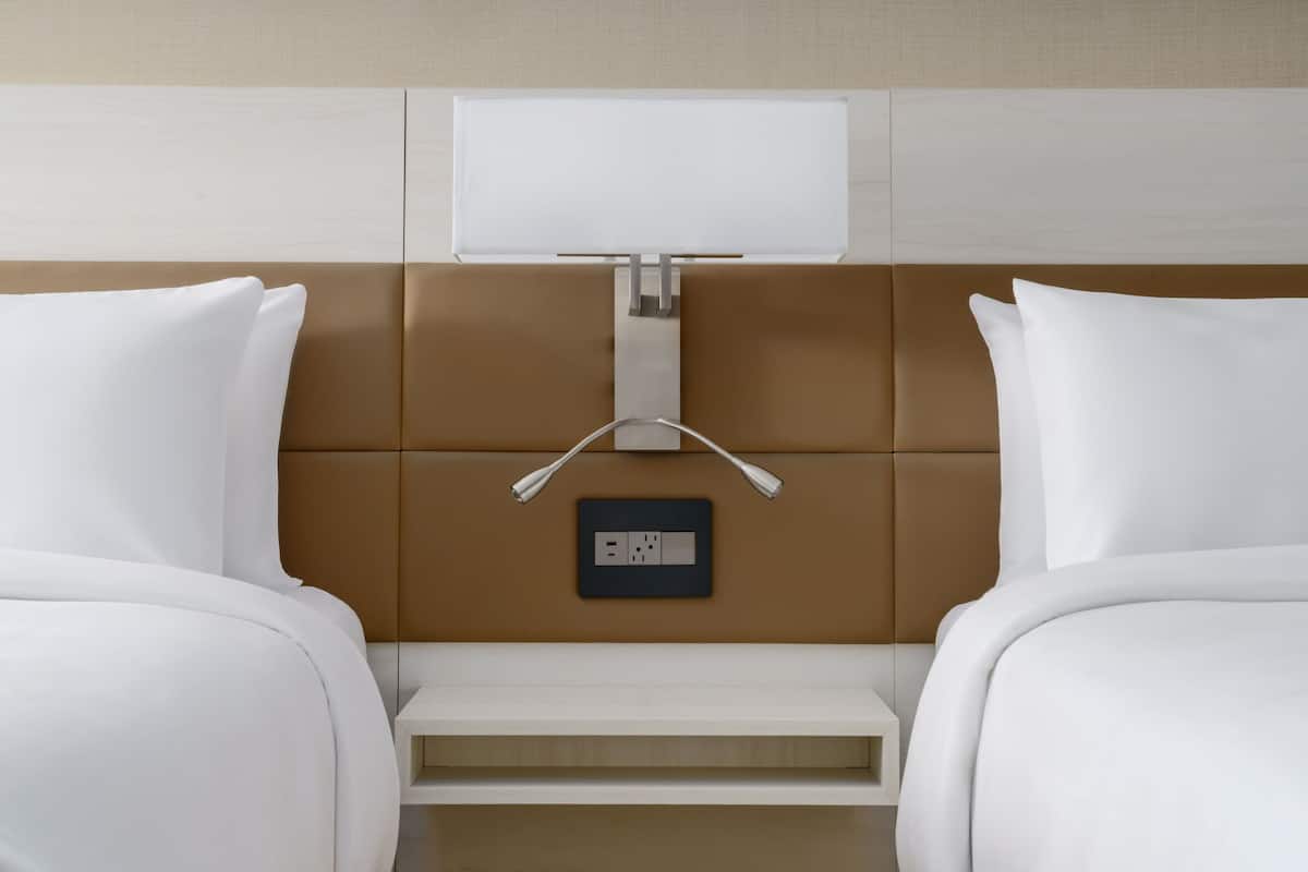 A hotel room with two beds, a bedside table, a wall-mounted TV, and a power outlet.