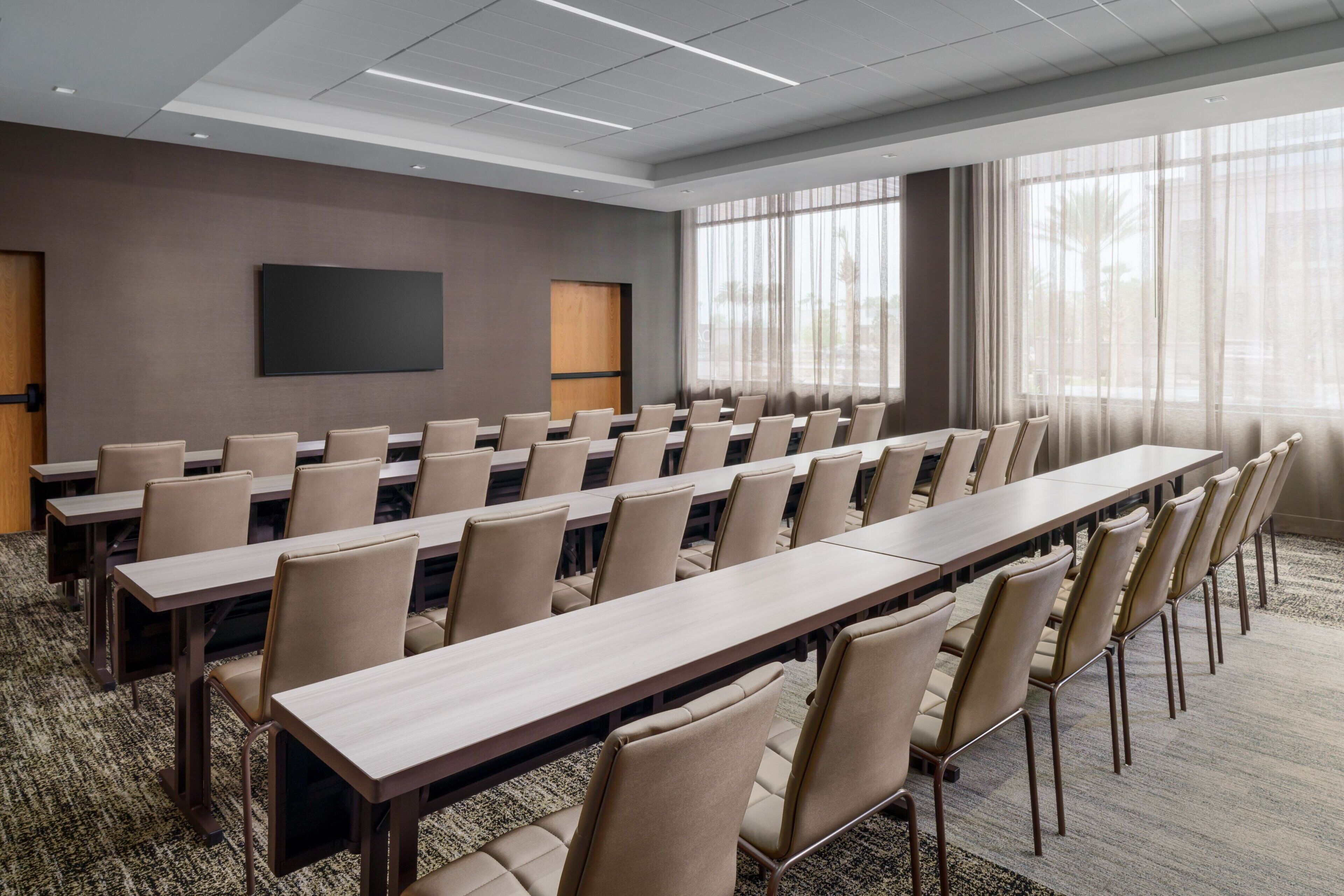 A conference room with long tables and chairs, a large screen, and a view of the outdoors through large windows.