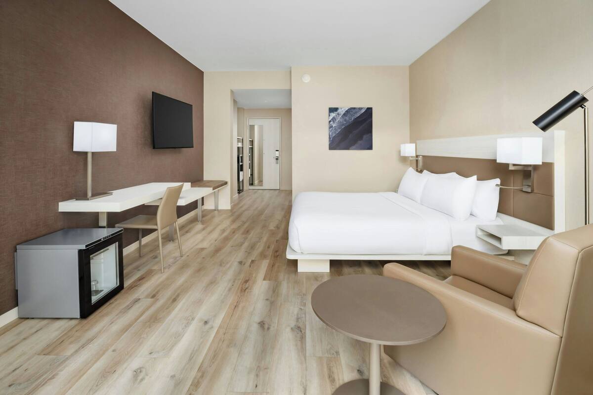 A hotel room with a bed, a desk, a chair, a small refrigerator, a bedside table, a lamp, and a wall-mounted TV.