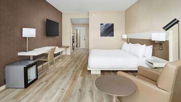 A hotel room with a bed, a desk, a chair, a small refrigerator, a bedside table, a lamp, and a wall-mounted TV.