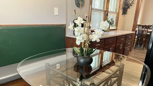 Una mesa de comedor con cubierta de vidrio, un jarrón con orquídeas blancas, armarios de madera y una ventana con persianas en el fondo.