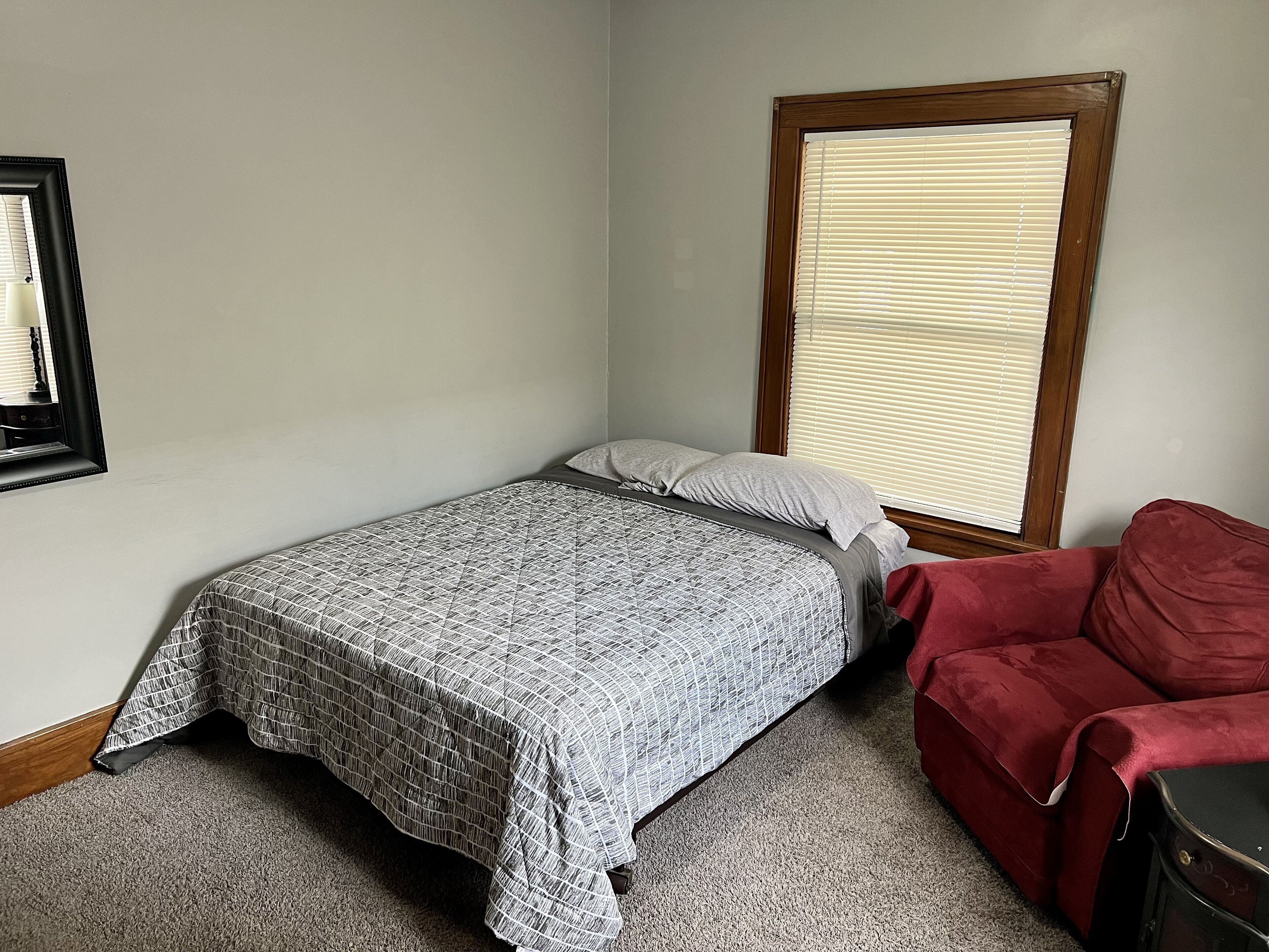 A small, neatly arranged bedroom with a single bed, a red chair, and a window with blinds.