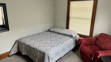 A small, neatly arranged bedroom with a single bed, a red chair, and a window with blinds.