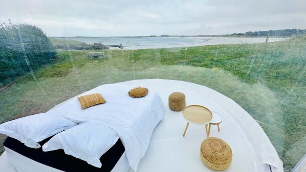 Een slaapkamer met een bed, kussens, een ronde tafel en een stoel, met uitzicht op een grasveld en een waterpartij.