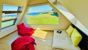 Een knus loft slaapkamer met een groot raam met uitzicht op een grasveld en de zee.