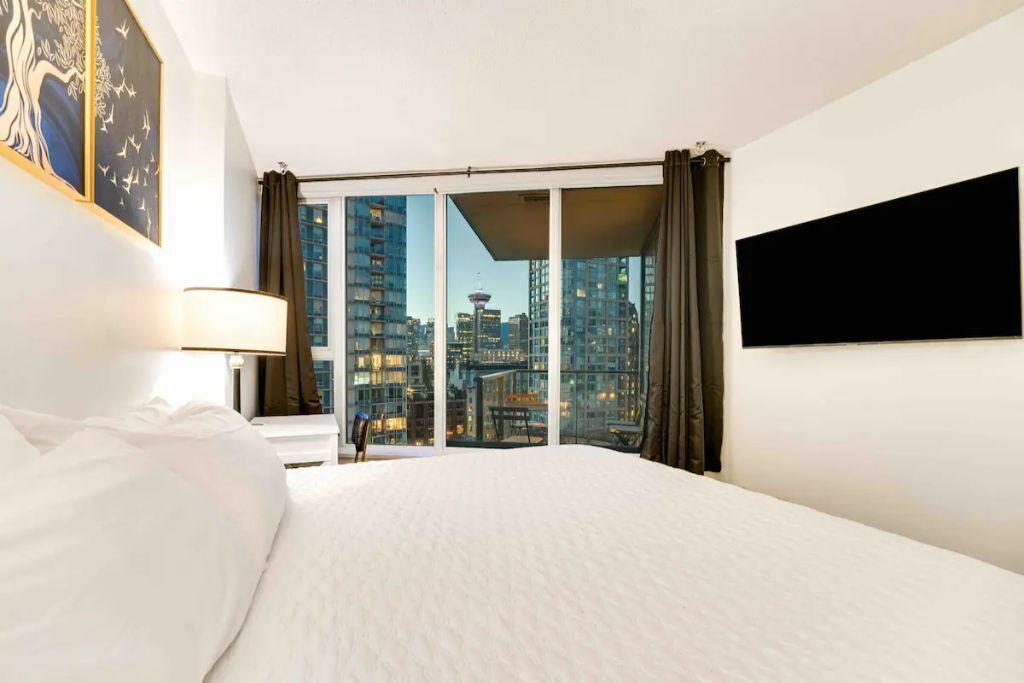 Une chambre d’hôtel avec un grand lit, une vue sur la ville grâce à de grandes fenêtres du sol au plafond, un téléviseur à écran plat fixé au mur et un tableau accroché au mur.