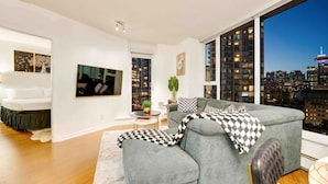 A modern living room with a grey sectional sofa, a flat-screen TV, and a large window offering a city view.