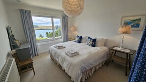 A bedroom with a large bed, two bedside tables, a window with a view of the sea, and a painting on the wall. - Fiddlers Green -  a holiday home that sleeps 12 guests  in 5 bedrooms (St Mawes)