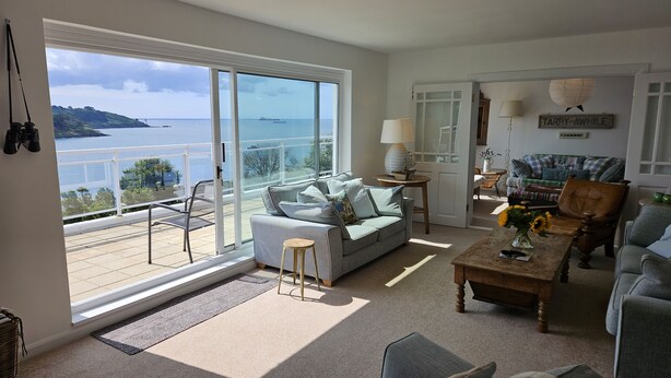 A spacious living room with a large sliding glass door leading to a balcony overlooking a scenic coastal view. - Fiddlers Green -  a holiday home that sleeps 12 guests  in 5 bedrooms (St Mawes)