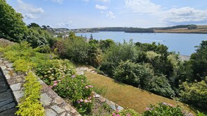 A garden with a stone pathway leading to a grassy area with pink flowers, overlooking a lake with boats. - Fiddlers Green -  a holiday home that sleeps 12 guests  in 5 bedrooms (St Mawes)