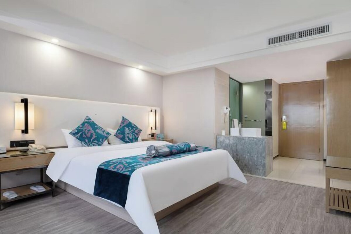 A hotel room with a large bed, a bedside table, a nightstand, a bathroom with a mirror, and a wooden door.