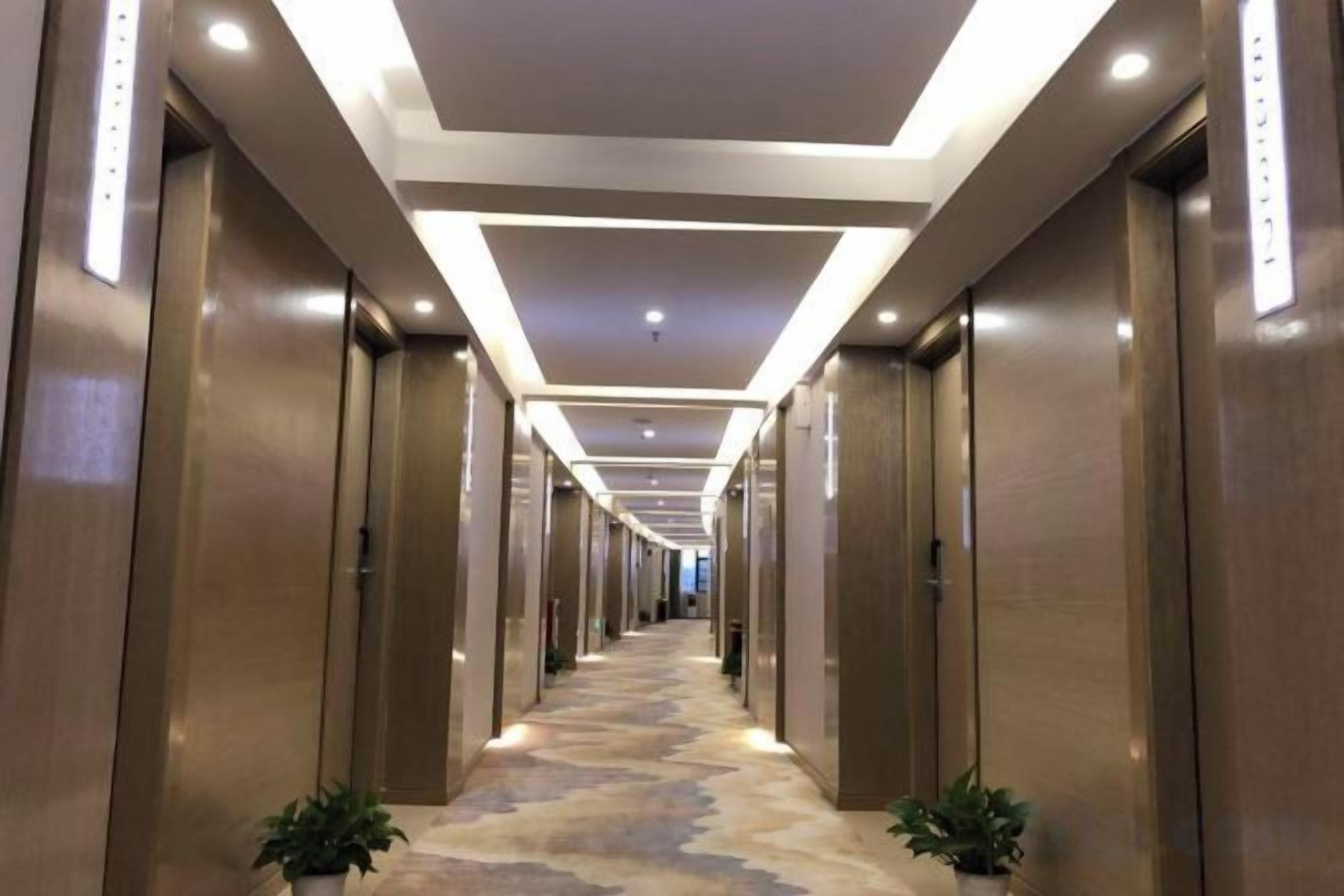 Corredor de hotel com piso revestido de carpete, portas de madeira e iluminação embutida.