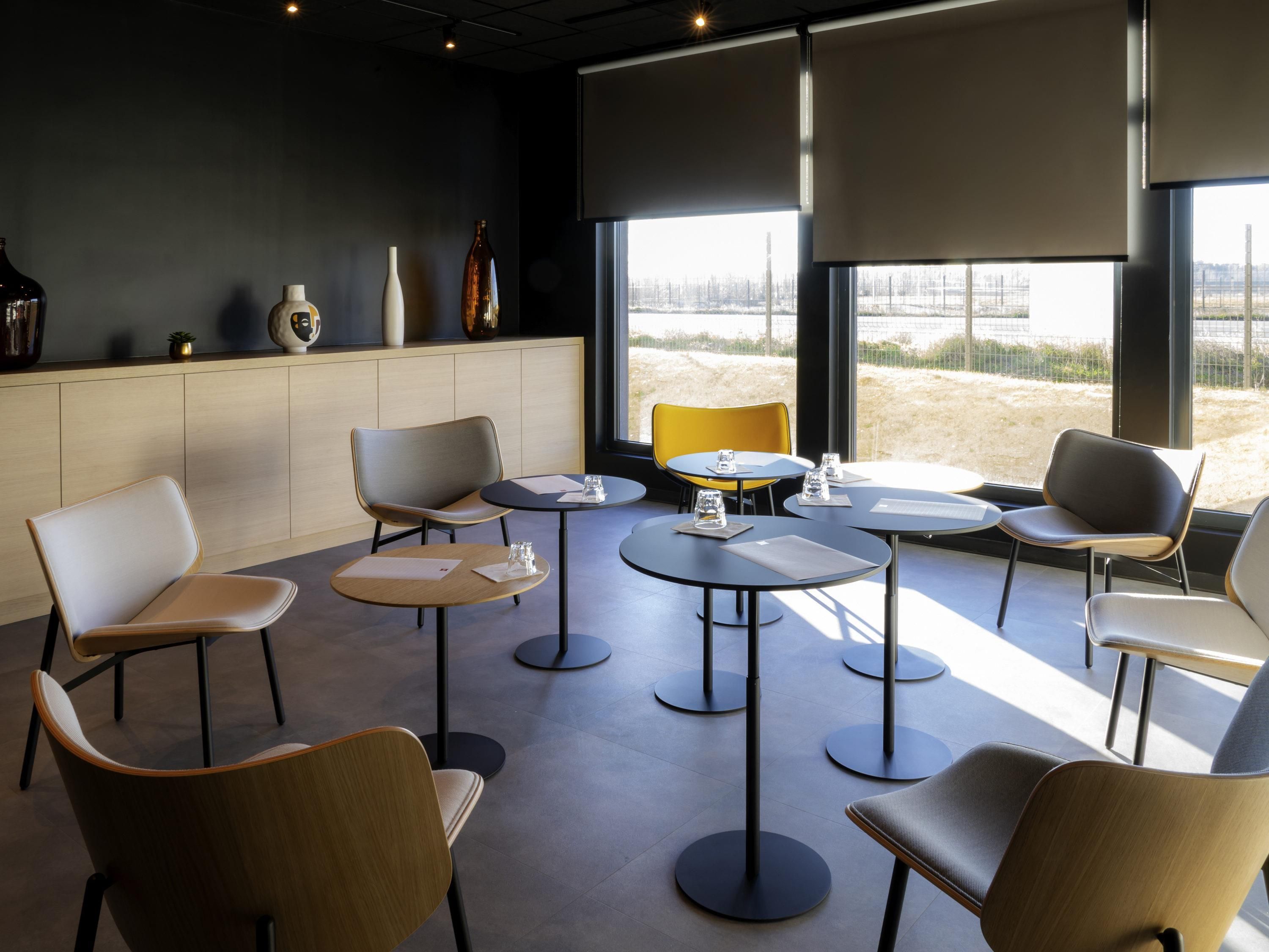 A modern interior with round tables and chairs, a wooden cabinet with decorative items, and large windows with a view of a field.