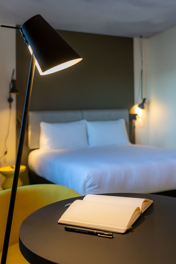Ein ordentlich bezogenes Bett mit weißen Leinentüchern, eine Nachttischlampe und ein kleiner Tisch mit offenem Notizbuch und Stift.