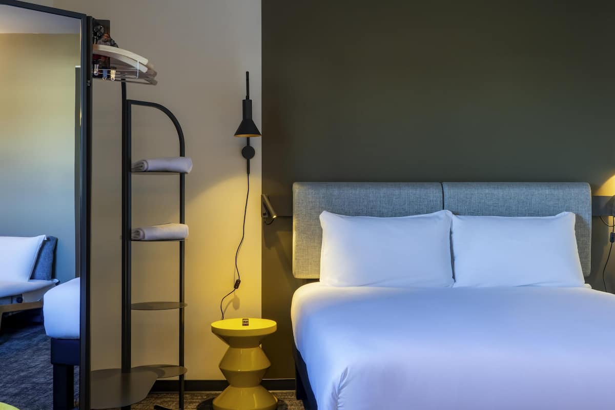Habitación de hotel con cama, mesita de noche, lámpara, estante con toallas y armario.