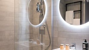 Une salle de bain moderne avec une douche à parois de verre, un lavabo blanc doté d’un distributeur de savon noir et un miroir avec un éclairage circulaire.