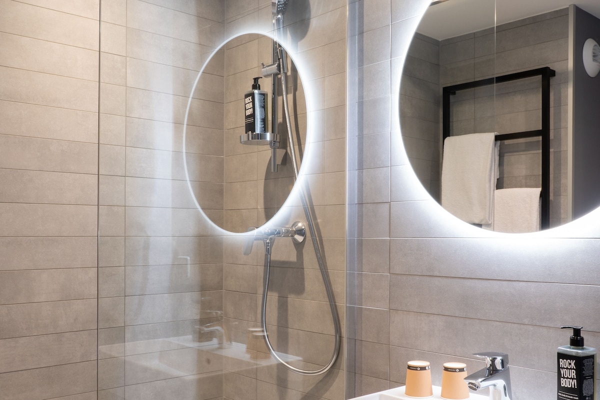 A modern bathroom with a glass-enclosed shower, a white sink with a black soap dispenser, and a mirror with a circular light.