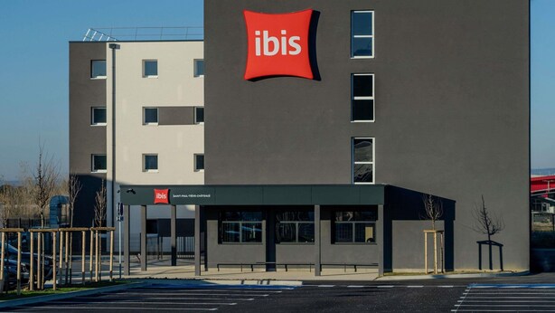 Un bâtiment moderne, facilement reconnaissable à son grand panneau rouge avec l’inscription « ibis », doté de nombreuses fenêtres et disposant d’un parking.