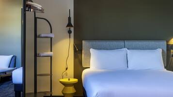 Habitación de hotel con cama, mesita de noche, lámpara, estante con toallas y armario.