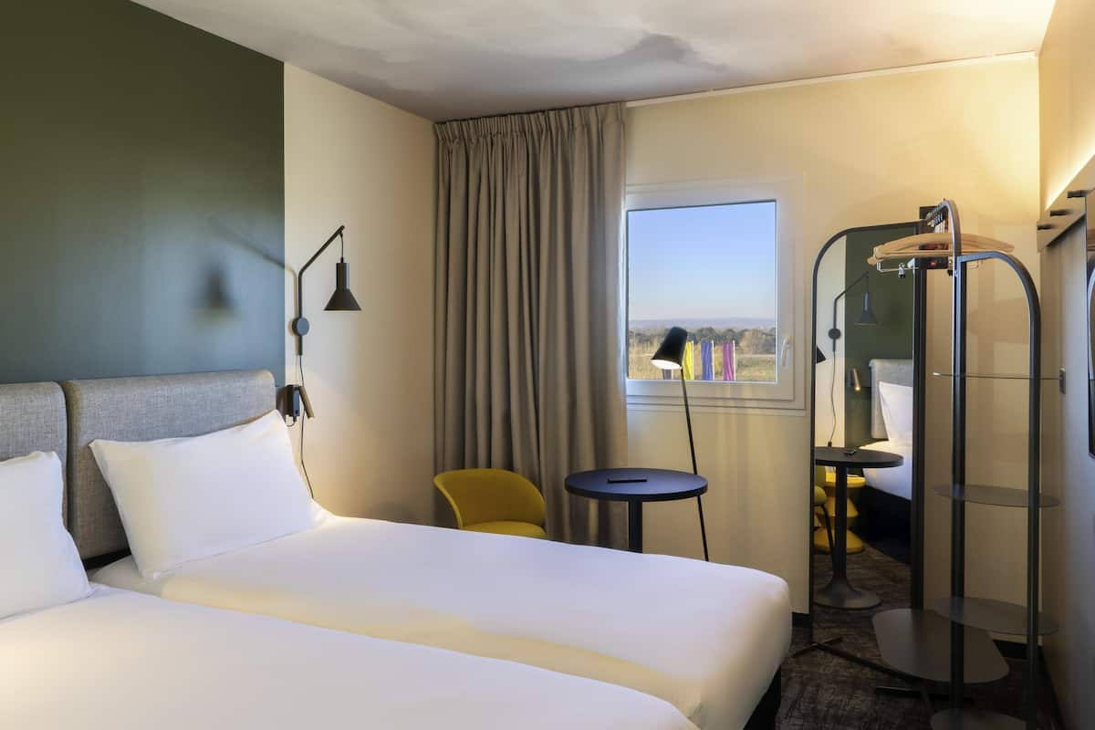 Habitación de hotel con dos camas, una mesita, una silla, ventana con cortinas y zona de ducha.