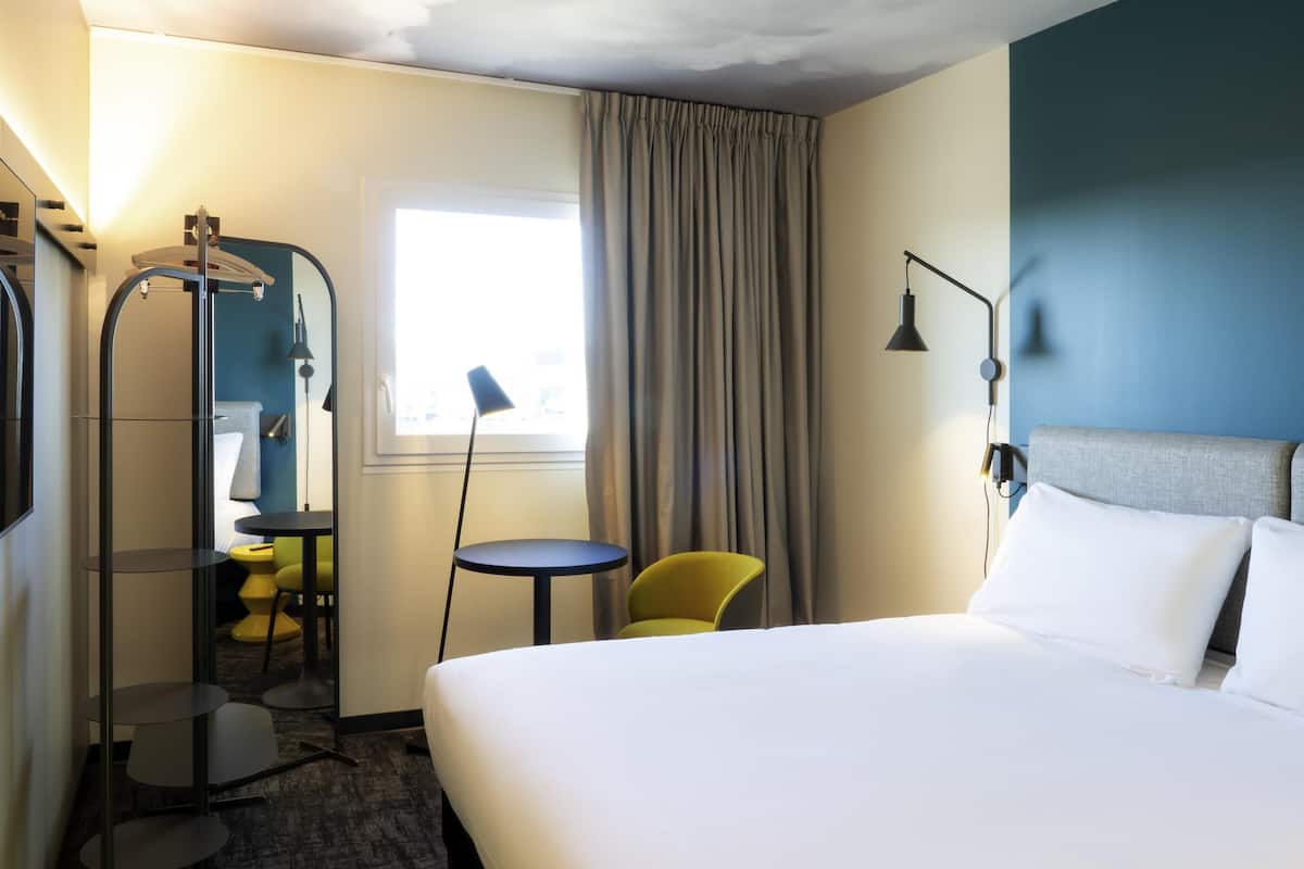 Habitación de hotel con cama, una silla, una mesita, un espejo y ventana con cortinas.