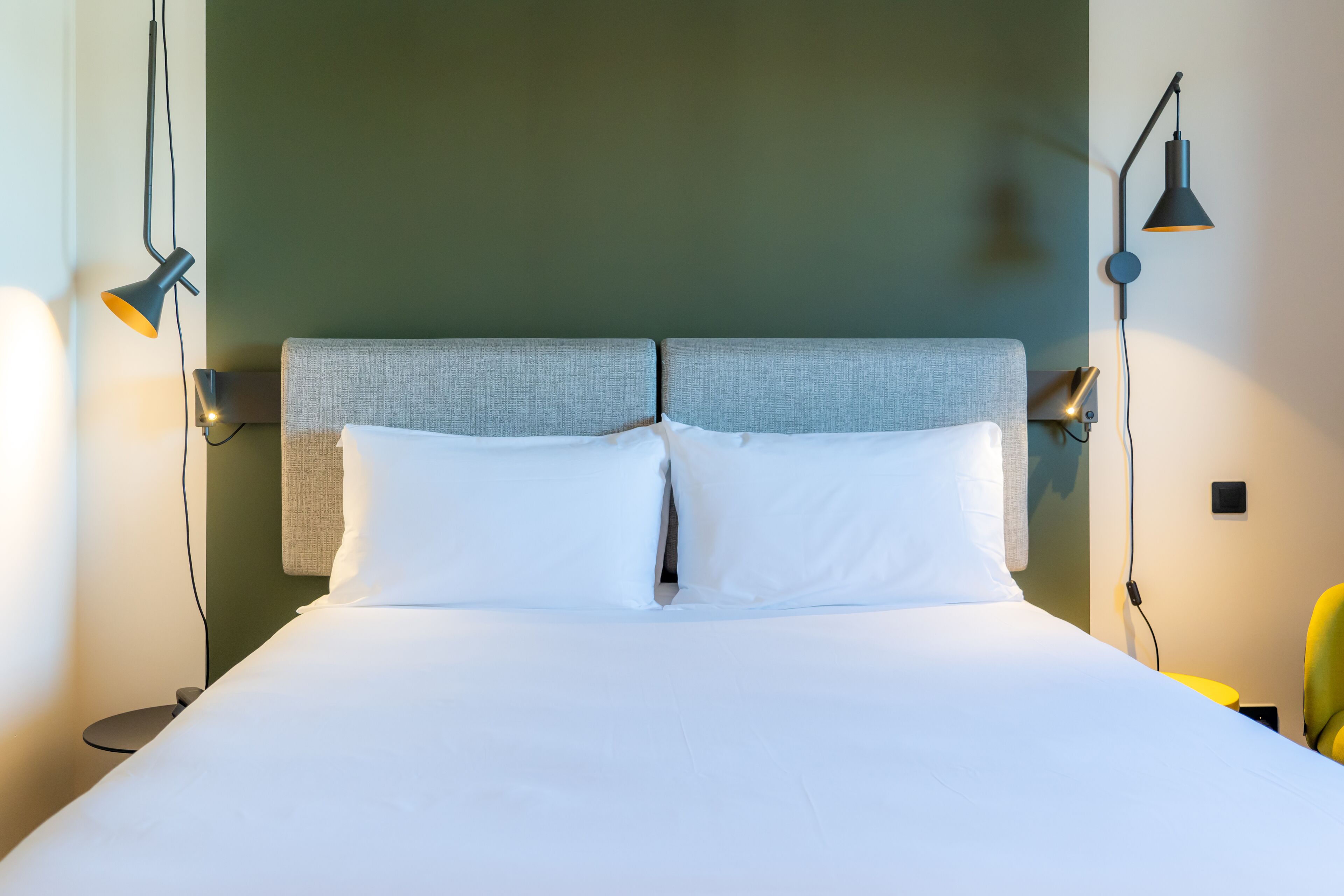 Una cama bien hecha con sábanas blancas, cabecera gris y dos lámparas fijadas a la pared a cada lado.
