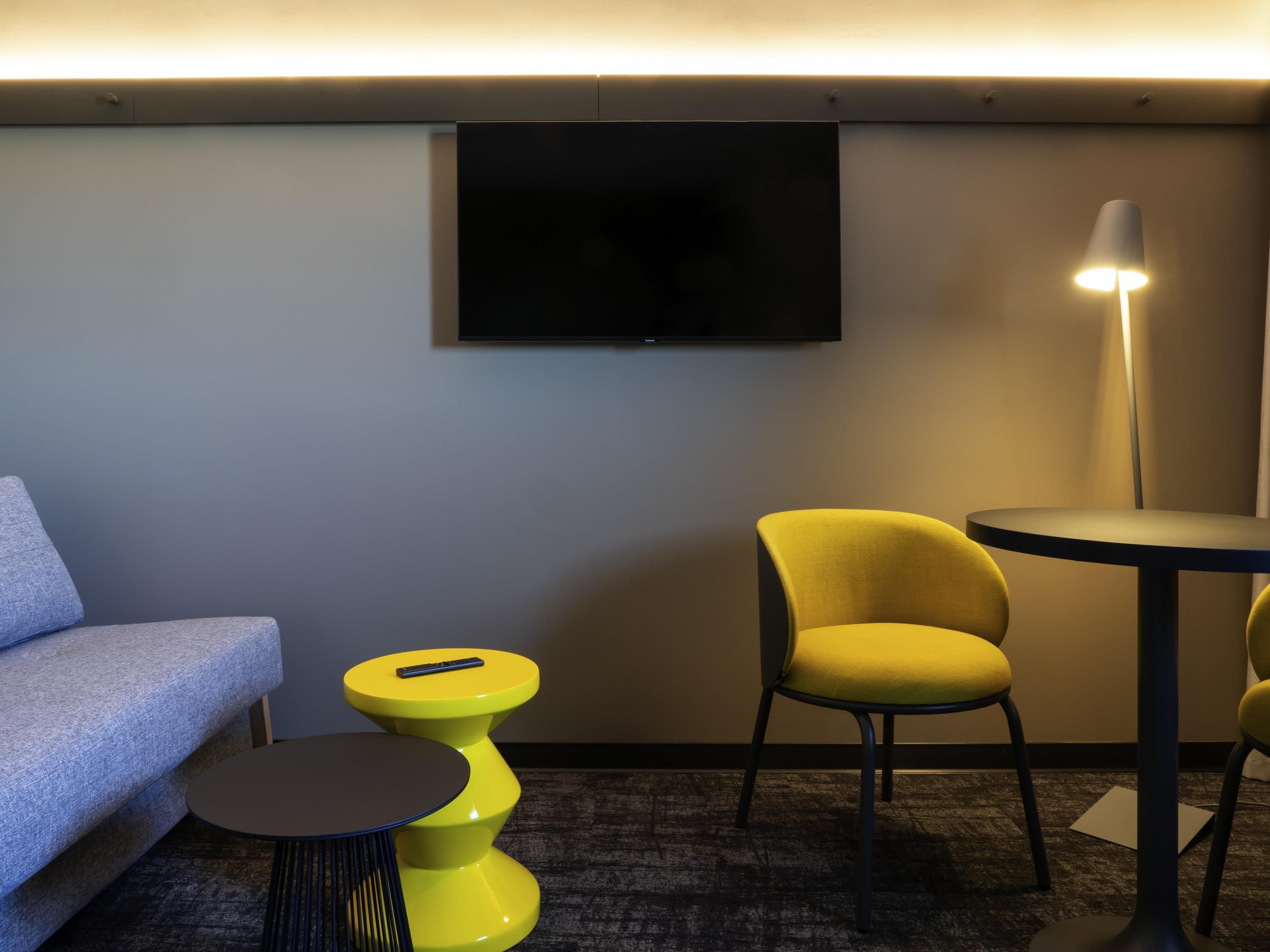 Ein modernes Hotelzimmer mit einer Couch, einem gelben Sessel, einem kleinen runden Tisch, einem an der Wand befestigten Fernseher und einer Stehlampe.