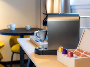 Una cafetera compacta sobre una mesa con una bandeja, dos tazas y una caja de cápsulas de café.