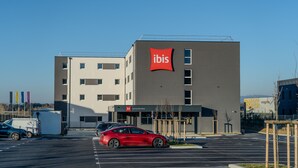 Un bâtiment moderne, orné d’une grande enseigne rouge « ibis », un parking où sont garées plusieurs voitures et un ciel d’un bleu éclatant.