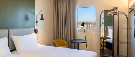 A hotel room with two beds, a small table, a chair, a window with curtains, and a shower area.