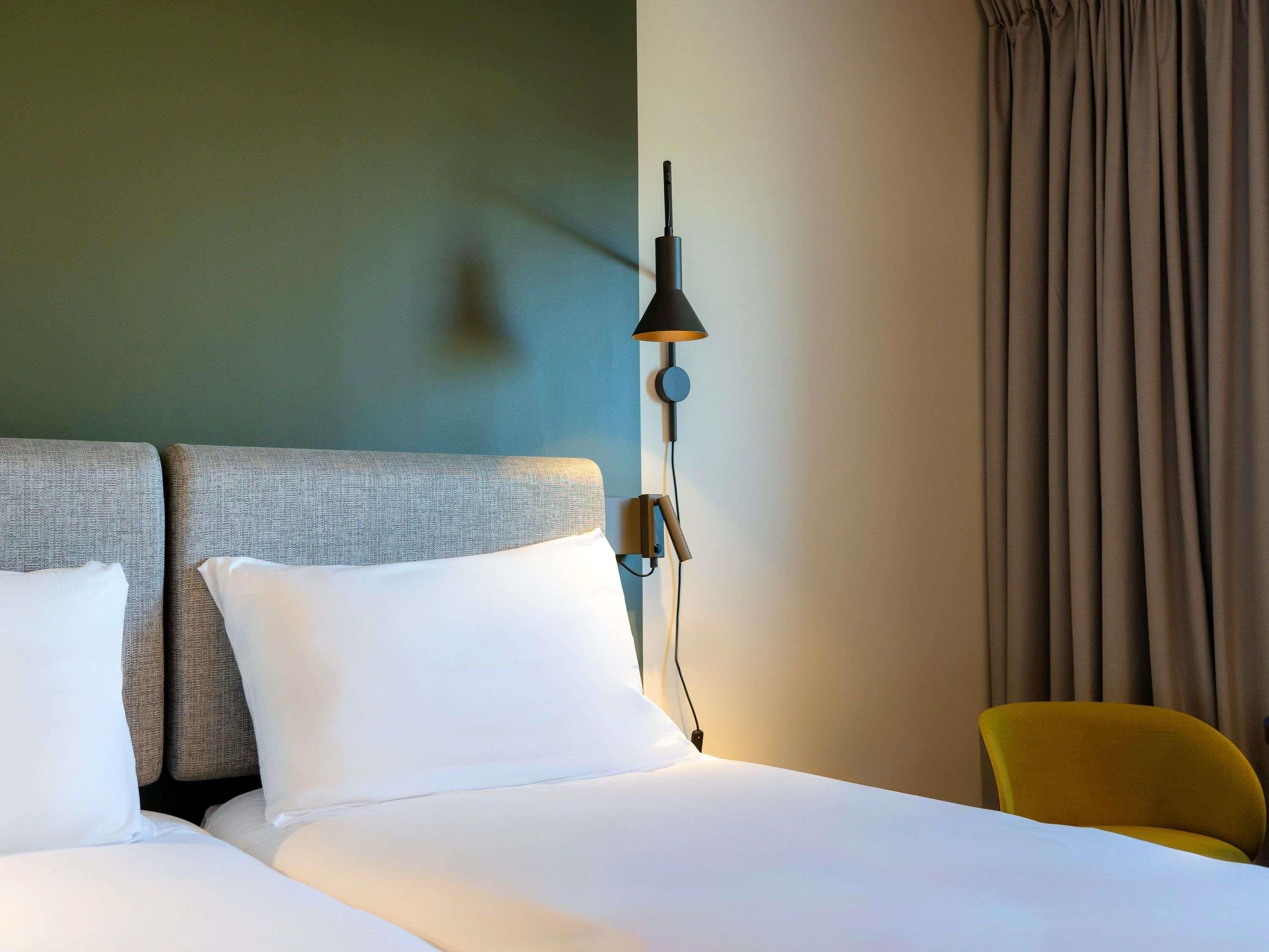 A hotel room with a bed, a nightstand, a lamp, a chair, and a curtain.