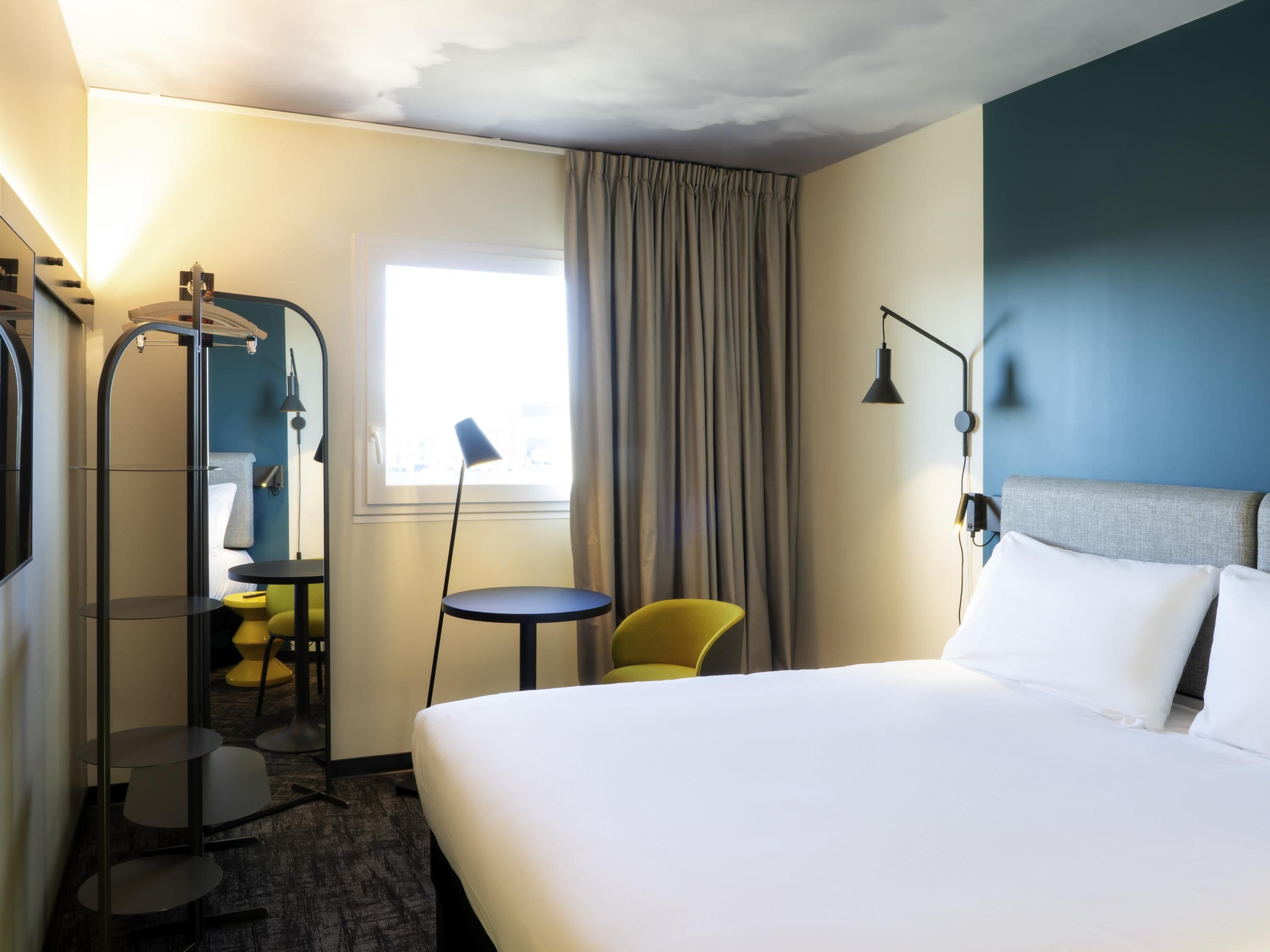 A hotel room with a bed, a chair, a small table, a mirror, and a window with curtains.